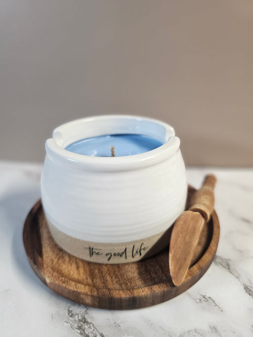 White ceramic pot filled with blueberry mornings candle. Has the good life writen on the front. Woth a wooden knife sitting next to it