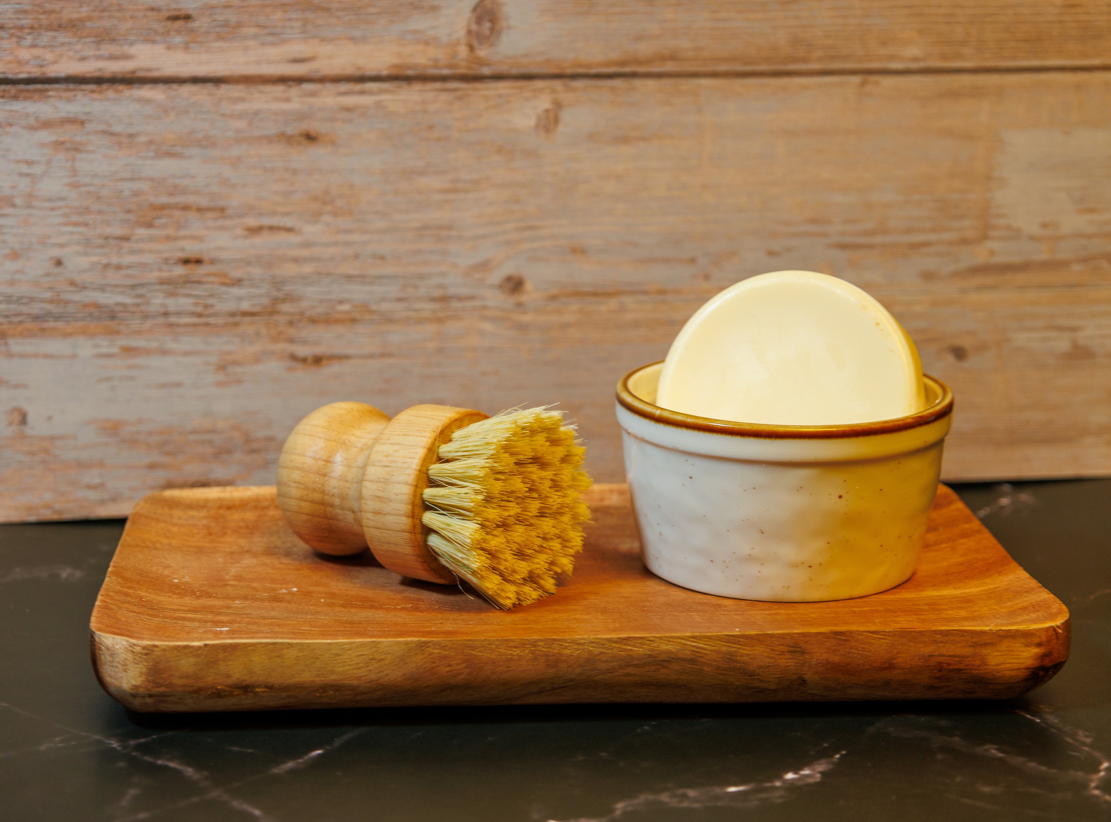 Citrus dish soap sitting in a cream colored ramekin on a wooden tray with a scrub brush laying next to it with a wooden background 