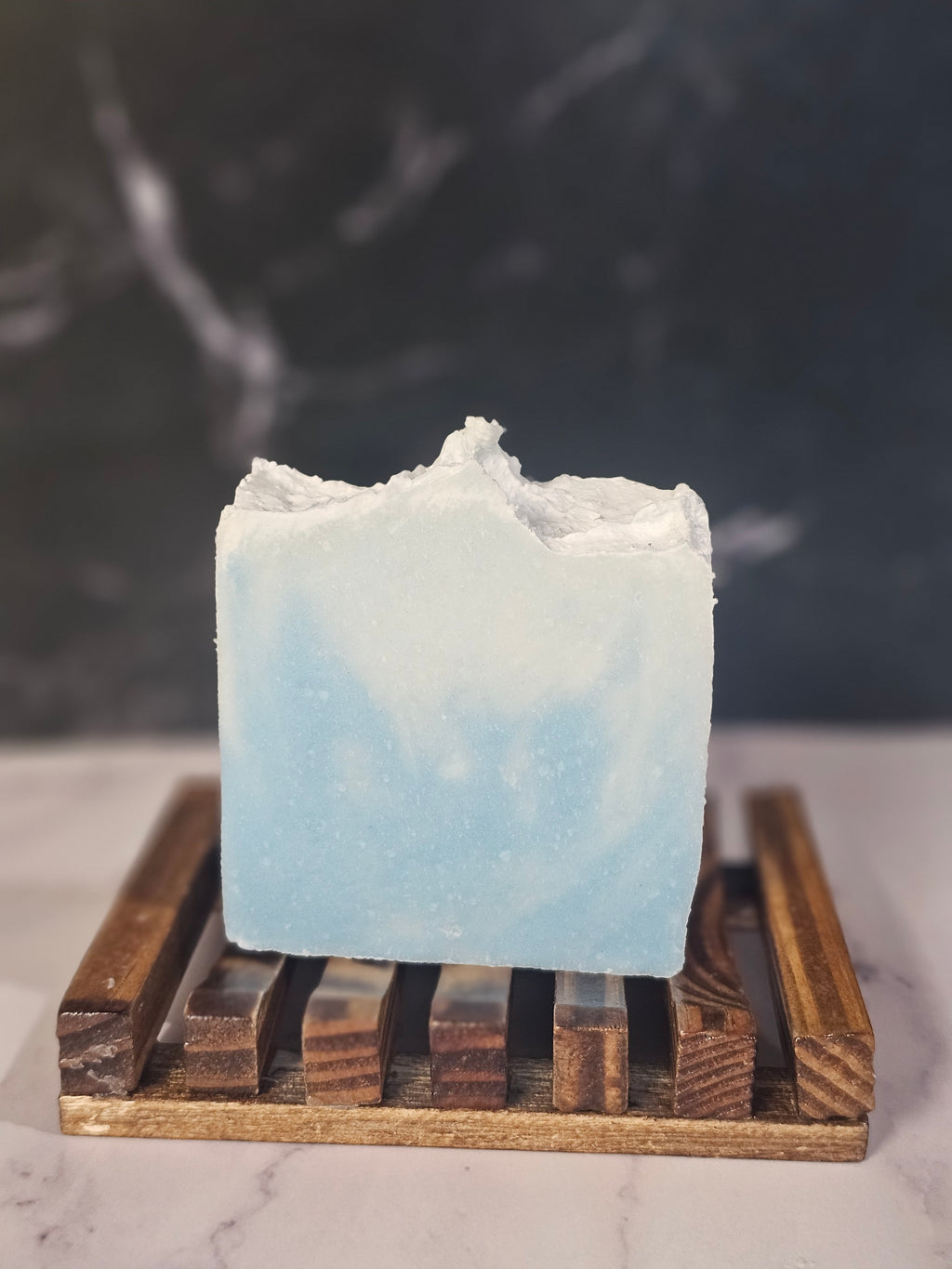 Bar of coastal rains soap on a wooden soap dish against a marble background