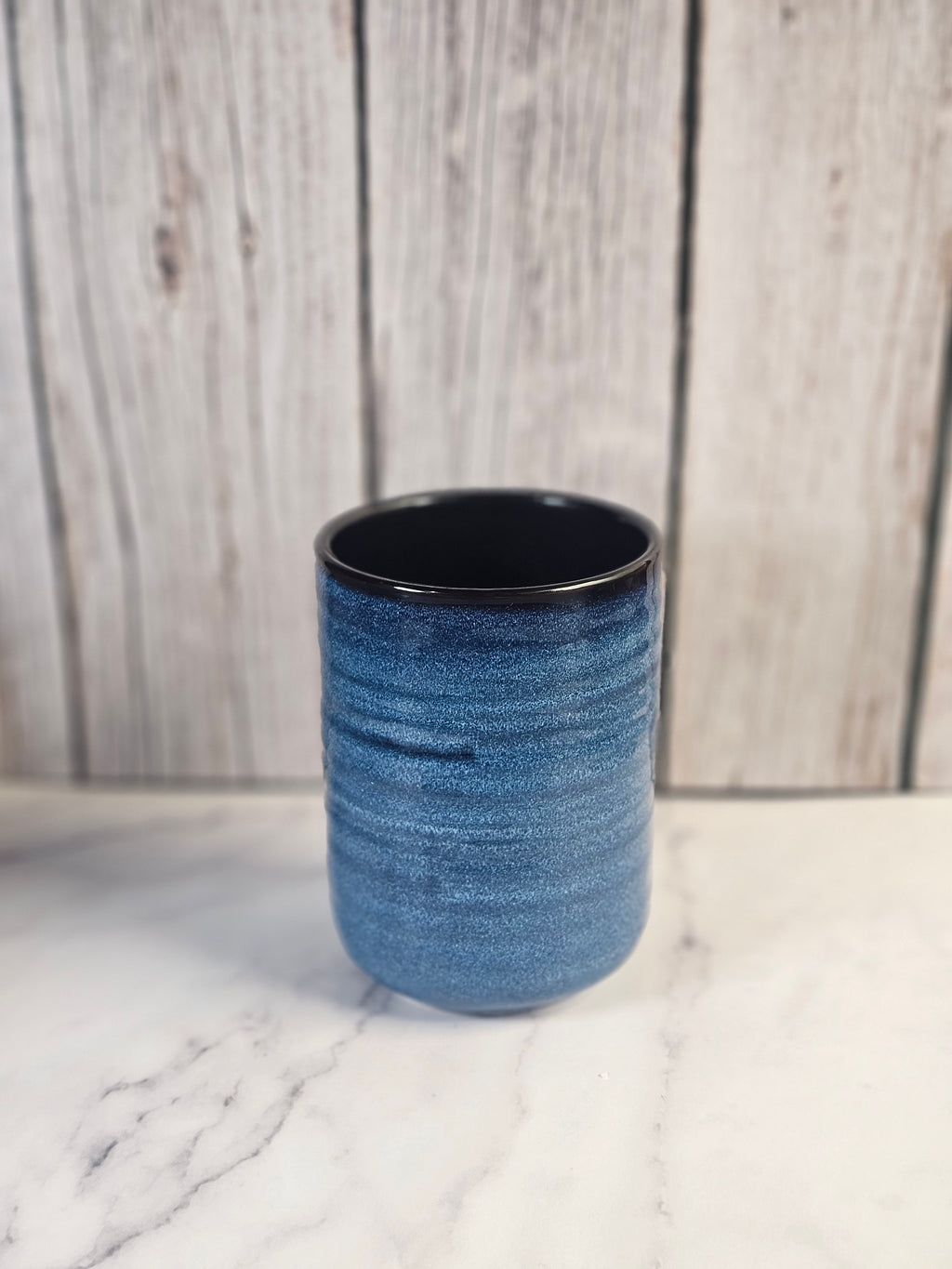 Small 24oz blue and black candle container sitting on a marble counter with a wooden background.