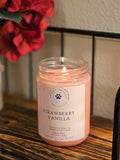 A strawberry vanilla candle in a clear glass jar sitting ona shelf next to some flowers