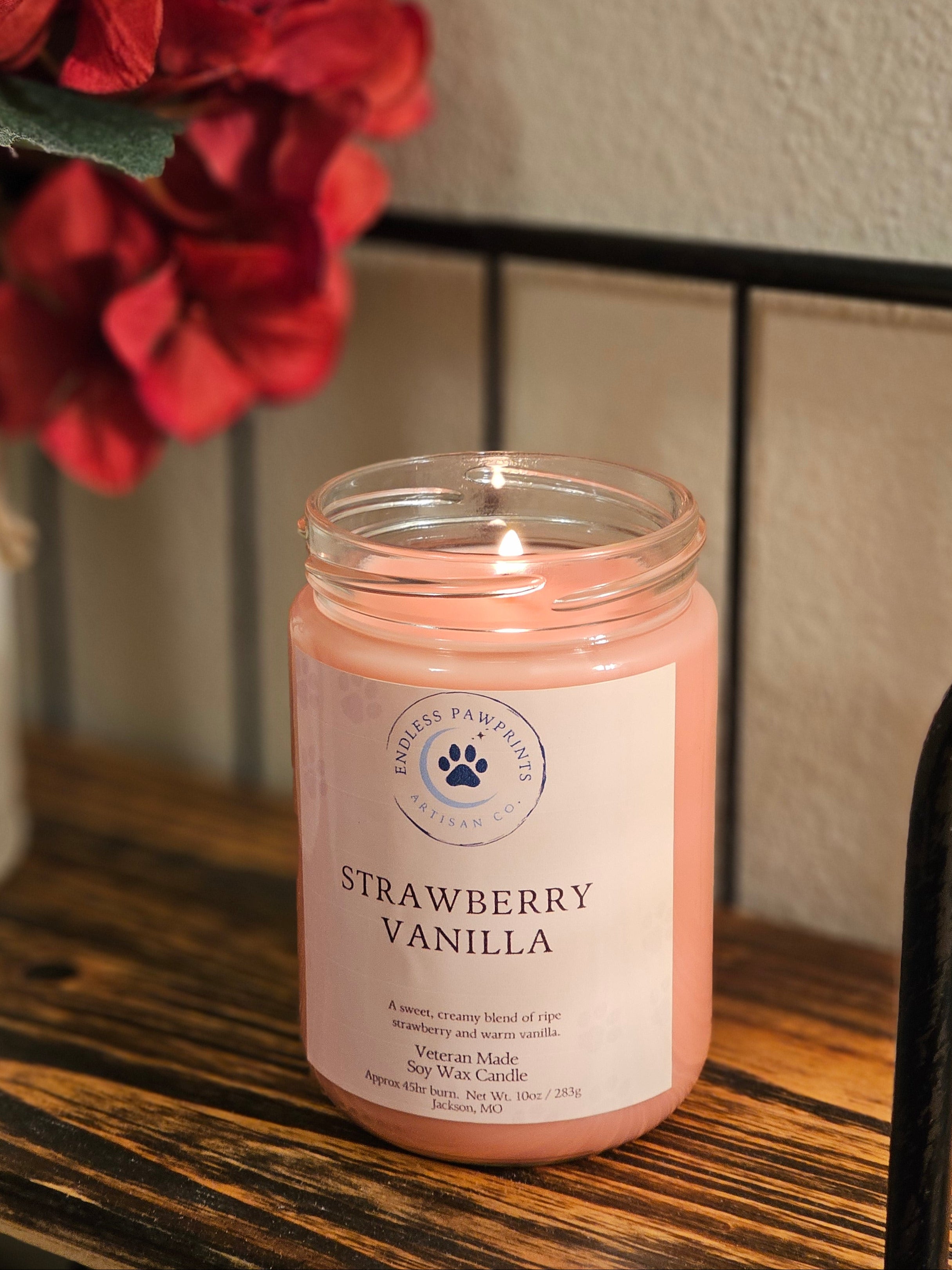 A strawberry vanilla candle in a clear glass jar sitting ona shelf next to some flowers