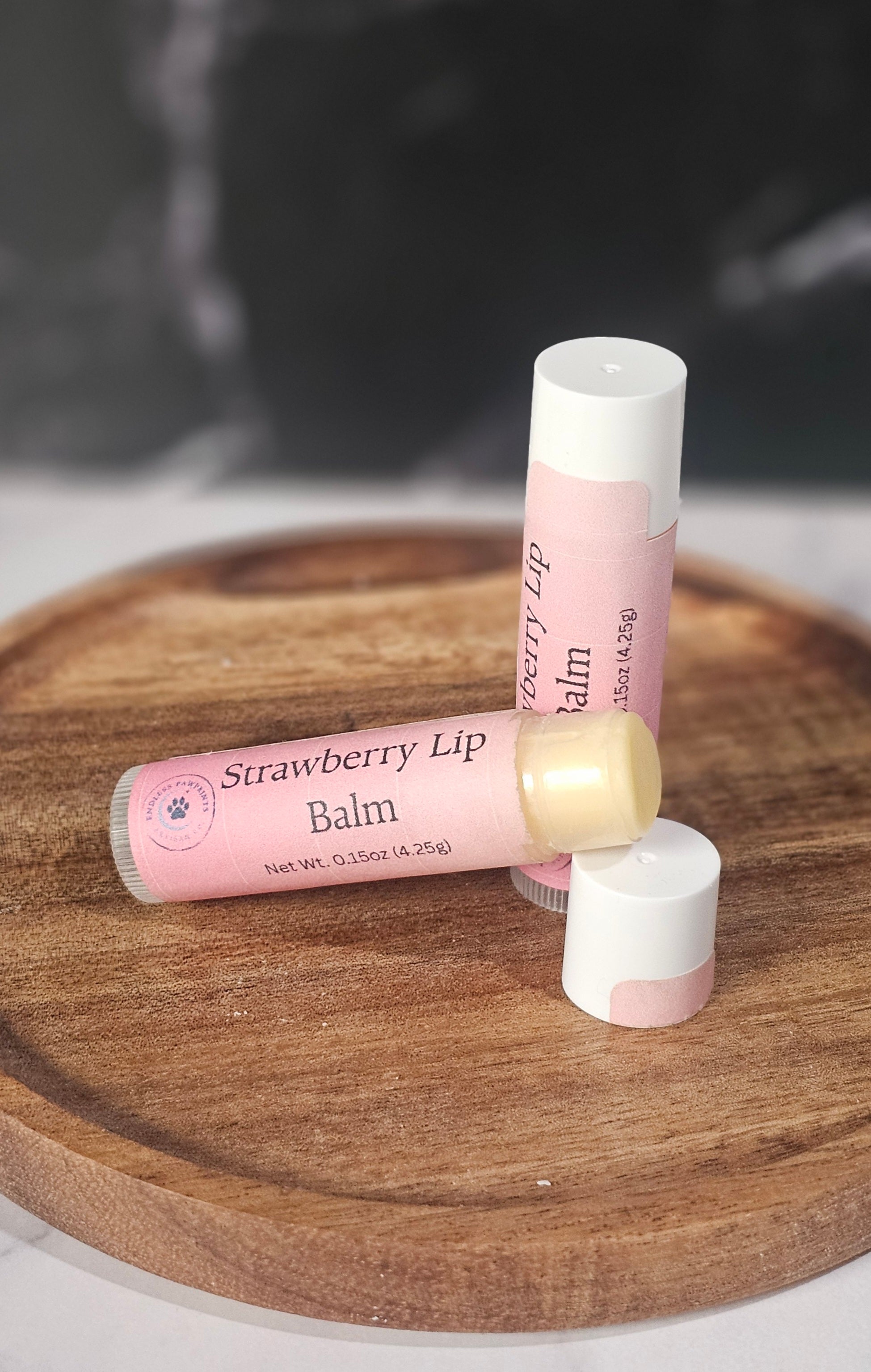 Two strawberry lip balm tubes on a wooden board with a dark background