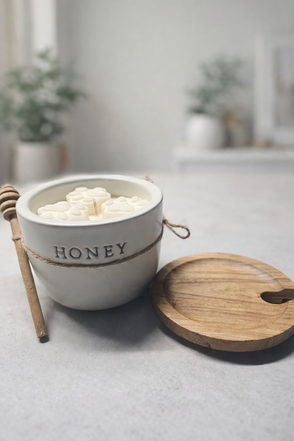 White ceramic honey pot with wooden lid and honey dipper on a light surface.