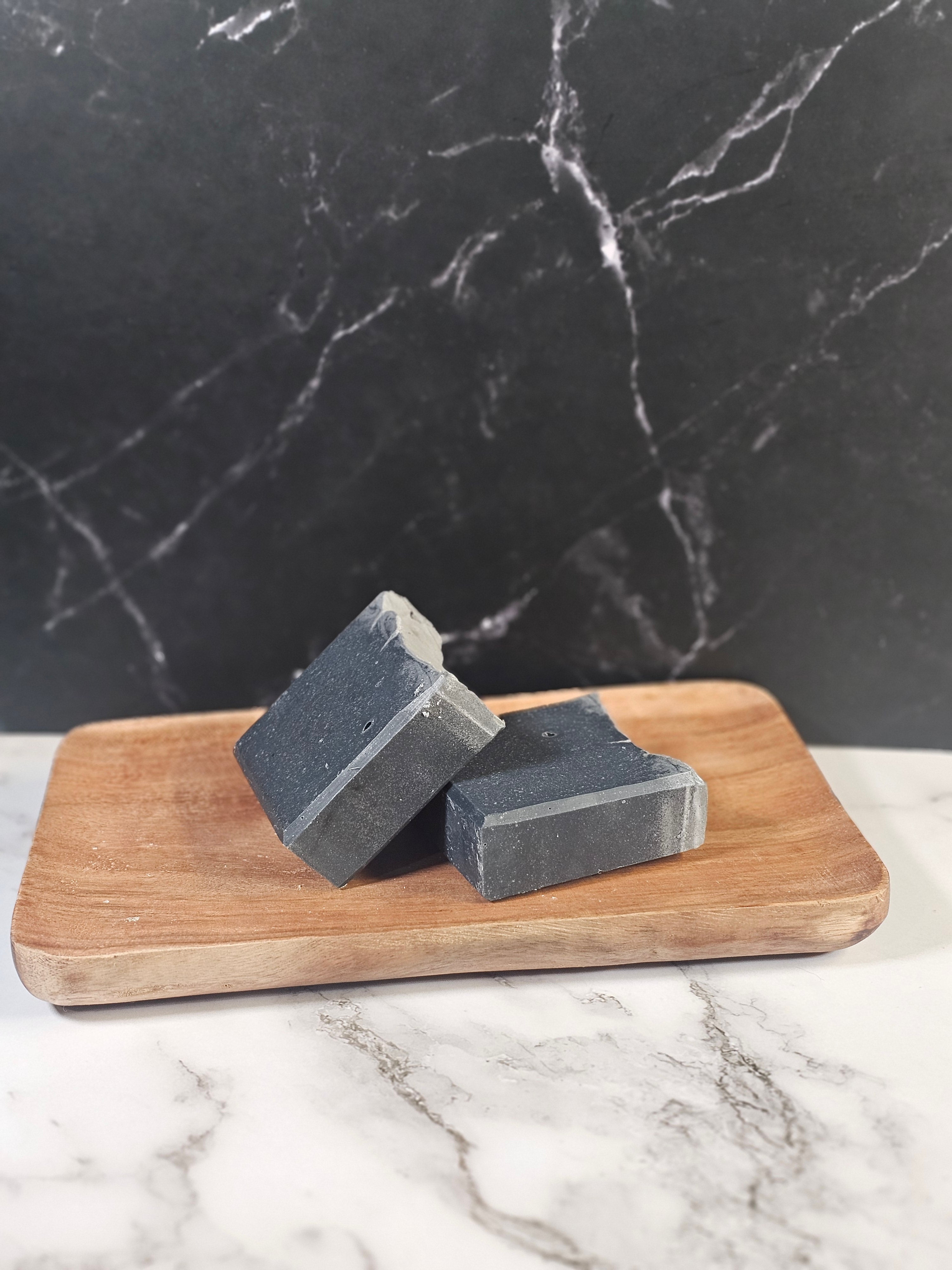 Two bars of charcoal soap on a wooden soap dish against a marble background