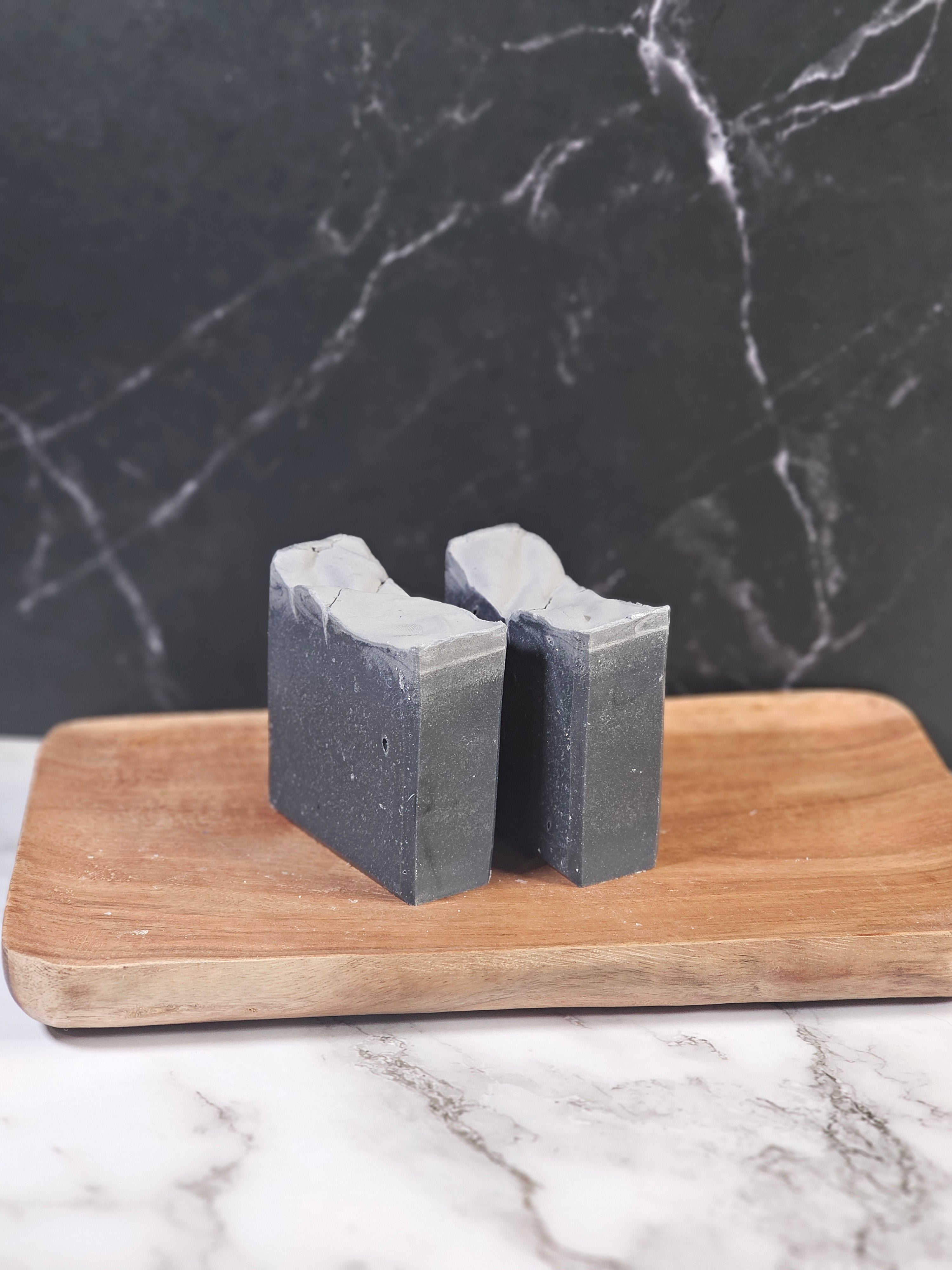 Two bars of black soap on a wooden cutting board against a marble background