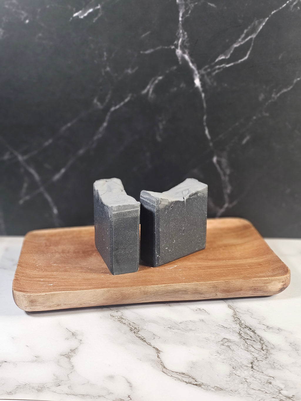 Two bars of midnight ash tallow soap sitting on a wooden board in front of a marble background