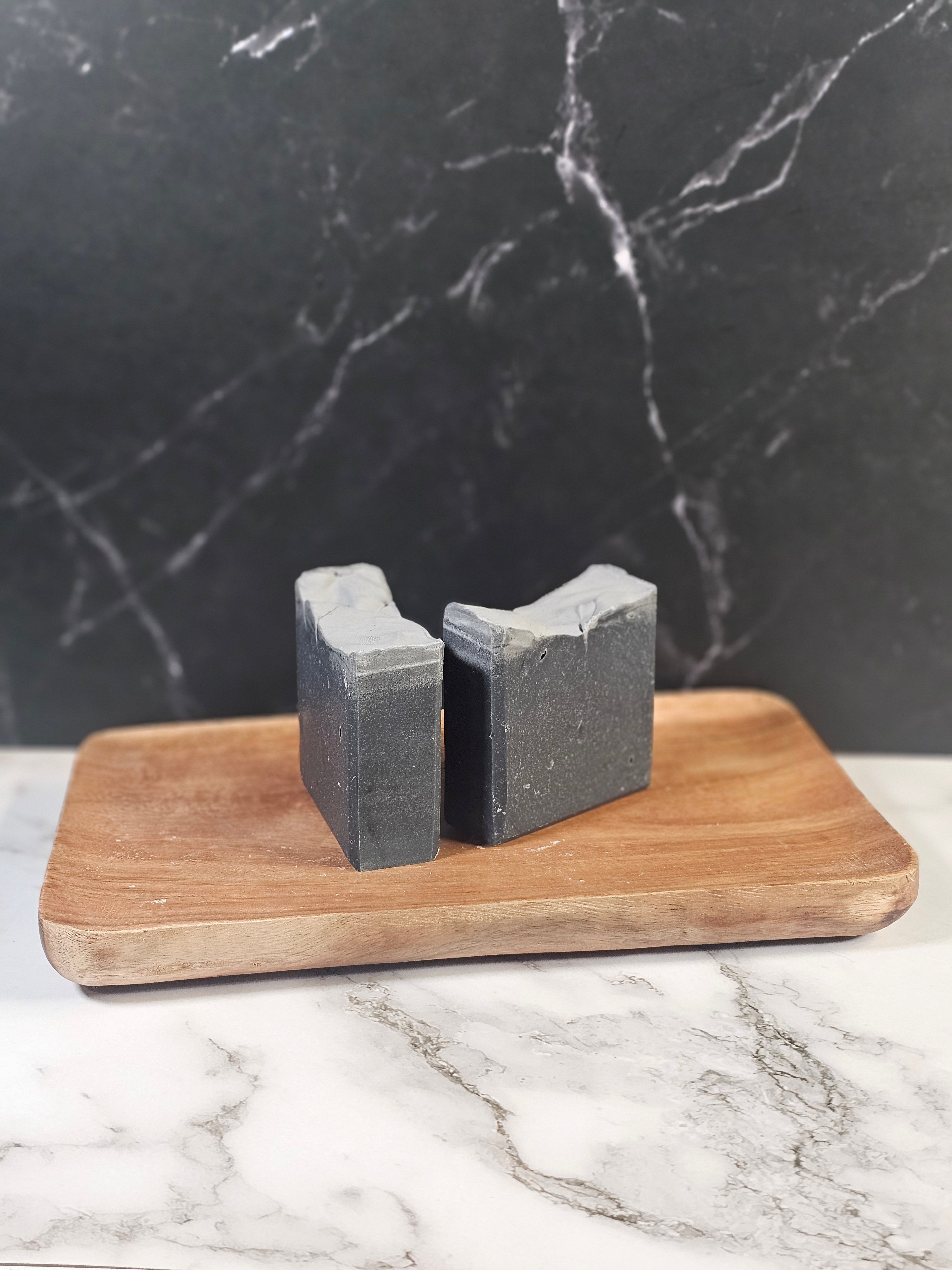 Two bars of midnight ash tallow soap sitting on a wooden board in front of a marble background
